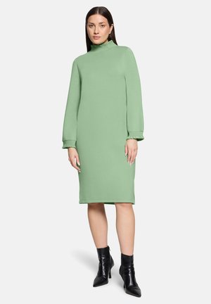 Femme portant une robe pastel verte longueur genou, à manches longues avec des poignets volantés, associée à des bottines noires à bouts pointus.