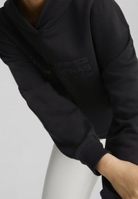 Sudadera con capucha negra, fabricada en una mezcla de algodón con una textura suave. Presenta puños acanalados y un sutil logotipo impreso en la parte frontal.