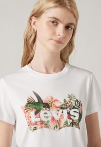 Camiseta blanca de algodón con un gráfico colorido de flores y un colibrí, con el texto audaz "Levi's" en un color contrastante en la parte frontal.