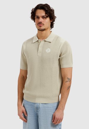 Man met krullend haar, gekleed in een beige korte mouw gebreid poloshirt met logo en lichtblauwe spijkerbroek, staande tegen een effen achtergrond.