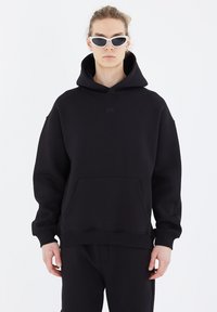 Black hooded sweatshirt made of soft fabric, featuring a front kangaroo pocket and subtle embossed logo. Model wears white sunglasses.