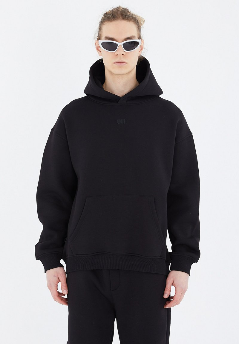 Black hooded sweatshirt made of soft fabric, featuring a front kangaroo pocket and subtle embossed logo. Model wears white sunglasses.