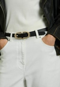 Svart läderbälte med gyllene oval spänne som bärs ovanpå vita denimbyxor med en slät yta och sidofickor. Ribbande vitt linne under en svart jacka.