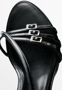 Sandale en cuir noir avec trois fines brides croisées sur le devant, chacune maintenue par une petite boucle rectangulaire argentée.