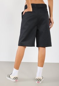 Shorts noirs ajustés à coupe droite, avec poches latérales et une texture tissée lisse, associés à des chaussettes blanches jusqu'à la cheville et des chaussures mocassins à carreaux.