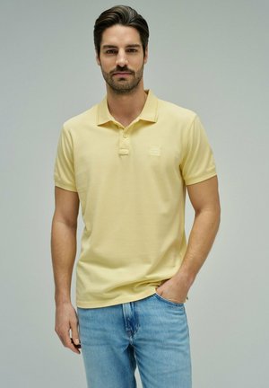 Hombre con una camisa tipo polo de manga corta de color amarillo pálido y jeans azul claro, de pie con una mano en el bolsillo frente a un fondo neutro.