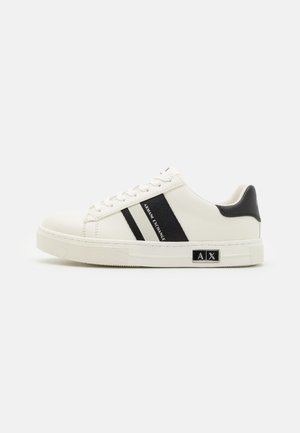 Sneakers in pelle bianca con accenti e strisce nere, punta rotonda, suola in gomma testurizzata e dettagli del logo sul lato.