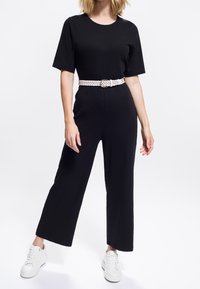 Kvinna klädd i en svart ribbad jumpsuit med korta ärmar, ett vitt flätat bälte och vita sneakers på en vit bakgrund.