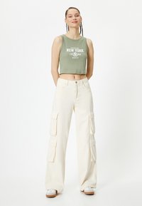 Top de tirantes verde cortado con texto blanco, combinado con pantalones cargo holgados de color crema que cuentan con grandes bolsillos laterales y un corte recto.