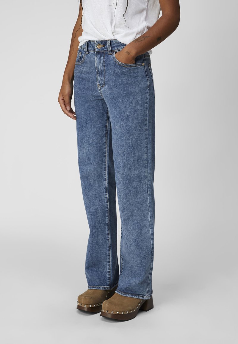 Blå denimjeans med høy midje og bred benprofil. Har klassisk fem-lommers design og beltehemper. Brukt med brune platåklosser.