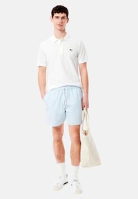 Camisa polo blanca con pequeño logo verde, pantalones de baño a rayas en tonos azul claro, calcetines blancos, zapatillas blancas y un bolso de lienzo crema.