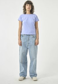 Helles lila kurzärmliges Oberteil, kombiniert mit locker sitzenden hellblauen Jeans aus Denim. Der Stoff wirkt weich und hat eine glatte Textur.