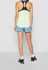 Jasnozielony sportowy top na ramiączkach z czarnym akcentem i logo z tyłu, w zestawieniu z niebieskimi szortami i czarnymi butami sportowymi, z białymi skarpetkami.