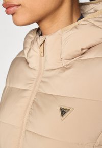 Beige dunjakke med hette, med glidelås og trekantet logo detalj. Glatt tekstur med quiltede seksjoner.