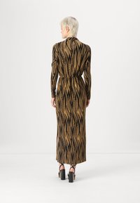 Maxi-Kleid aus einem strukturierten, goldenen Stoff mit schwarzen Zickzack-Mustern, langärmlig, mit tailliertem Schnitt und seitlichen Schlitzen. Schwarze Plateau-Highheels.