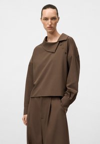 Top corto oversized marrone con un grande colletto e tre bottoni decorativi sulla spalla. Tessuto morbido e liscio con una vestibilità rilassata.