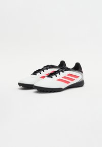 Weiße Sportschuhe mit schwarzen Akzenten, versehen mit drei roten Streifen, einer strukturierten Oberfläche und einer Gummisohle, die für Kunstrasen geeignet ist.