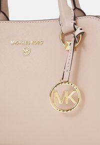 Sac à main beige Michael Kors avec lettres du logo dorées et un charm rond doré MK suspendu à la poignée.