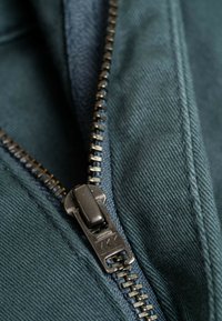 Detalle de prenda con cremallera que presenta un tejido en un tono verde azulado oscuro y una cremallera de metal VKK con dientes visibles y costura. La textura es suave y duradera.