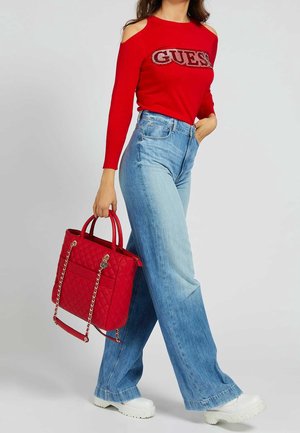 Femme portant un pull rouge à épaules dénudées avec le texte « GUESS », un jean bleu à jambes larges, des chaussures blanches, et tenant un sac à main rouge matelassé avec des anses-chaînes.