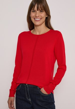 Femme souriante portant un haut à manches longues rouge vif et un jean bleu foncé, une main dans la poche, sur un fond uni.