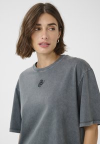 Tricou oversize din bumbac gri, cu guler rotund, mâneci scurte și un mic logo brodat negru pe piept. Textură moale.