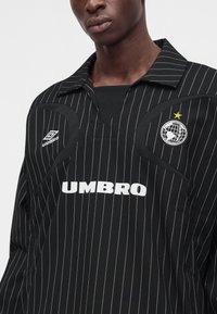 Мъж, носещ черен спортен екип Umbro на тънки райета с бели лога и глобус с жълта звезда на гърдите.