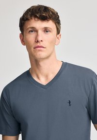 Grå V-ringad T-shirt tillverkad av lätt material med en liten mörk broderad logotyp på vänster bröst. Slät yta och avslappnad passform.