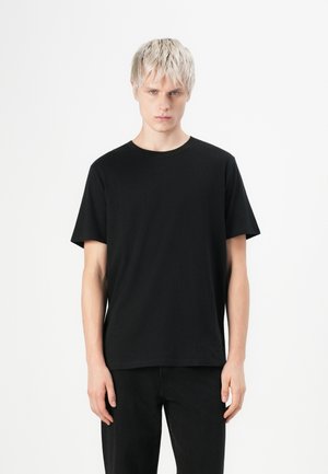 T-shirt en coton noir avec un col rond, des manches courtes et une coupe décontractée, portée avec un pantalon noir assorti sur un fond blanc uni.