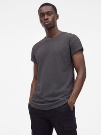 Young man standing with hands in pockets wearing a dark grey rolled-sleeve t-shirt and black cargo pants against a plain background.