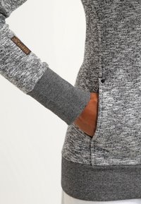 Main droite dans la poche d'un pull gris chiné avec poignets et ourlet côtelés gris foncé, peau de ton marron, et étiquette en tissu sur la manche indiquant "RAGEWEAR."