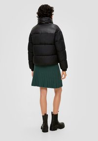 Schwarze Daunenjacke mit hohem Kragen, kombiniert mit einem grünen Faltenrock. Schwarze Stiefel mit dicken Sohlen runden das Outfit ab.