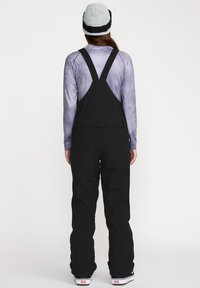 Salopette noire avec des bretelles réglables, grandes poches latérales et arrière, accompagnée d'un haut à manches longues violet et d'un bonnet en tricot gris.