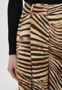 Záhybované nohavice s béžovým a čiernym zebra vzorom, hladkou textúrou a detailnou pružnou páskou, viditeľným logom na látke.