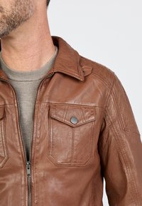 Braune Lederjacke mit einem Reißverschluss, zwei Brusttaschen, gepolsterten Schulterakzenten und einem Kragendesign. Weicher Stoff und taillierte Passform.