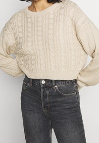 Pull beige en maille torsadée avec ourlet côtelé et coupe oversize, associé à un jean denim foncé taille haute avec poches avant et fermeture par bouton.