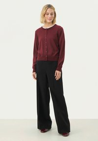 Burgunderfarbenen Cardigan mit Rundhalsausschnitt, ausgestattet mit einer Knopfleiste und langen Ärmeln, kombiniert mit schwarzen weit geschnittenen Hosen und kastanienbraunen Schuhen.