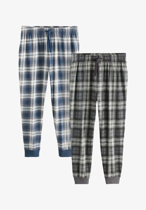 Homme torse nu avec des tatouages, portant un pantalon de pyjama à carreaux bleu et blanc, debout pieds nus sur un tapis dans une chambre avec un lit et une table de chevet.