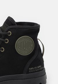 Botín de tobillo de lona negra con suela de goma, parche con logo texturizado y acentos verdes. Incluye cierre con cordones y detalle de coordenadas impresas.