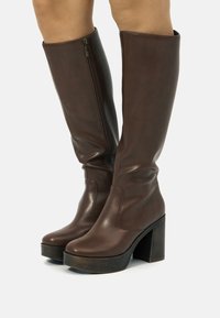 Botas marrones hasta la rodilla hechas de material sintético suave, con cremallera lateral, tacones gruesos y apilados, y un diseño de punta redonda.