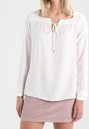 White long-sleeve blouse with a round neckline, tie front detail, and light, textured fabric. Paired with a pink denim skirt.