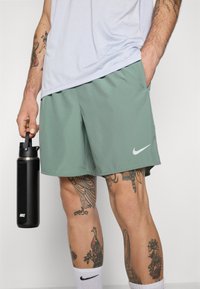 Pantalones cortos deportivos verdes hechos de material ligero, con un logo de Nike en blanco. Sostiene una botella de agua negra en una mano.