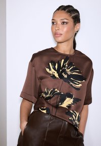 Blusa marrone in raso con maniche corte, caratterizzata da una grande stampa floreale nera e gialla. Tessuto liscio con vestibilità ampia e spacchi laterali.