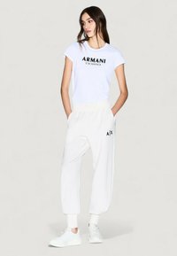 Camiseta blanca con el logo "ARMANI EXCHANGE" en negro, combinada con joggers blancos holgados que presentan un logo "A/X". Ambos artículos están hechos de un tejido suave.