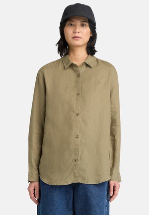 Kobieta w ciemnej czapce, oliwkowej koszuli na guziki z długim rękawem i niebieskich dżinsach, stojąca na tle gładkiego jasnego tła.