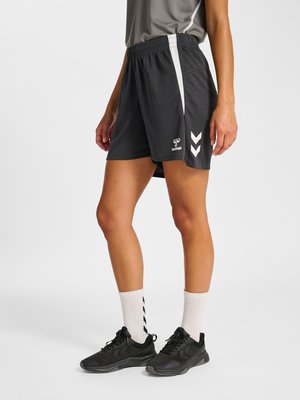 Mujer que lleva pantalones cortos deportivos negros con rayas blancas, calcetines blancos y zapatillas negras para correr, de pie contra un fondo liso.