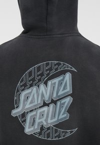 Hanorac negru cu un design grafic pe spate. Designul include textul "SANTA CRUZ" în albastru, cu modele circulare și linii.