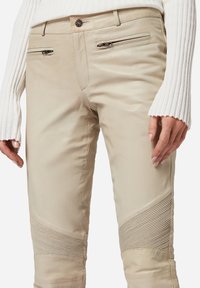 Beige Lederhosen mit glatter Textur, ausgestattet mit schrägen Reißverschlusstaschen und gerippten Akzenten an den Oberschenkeln. Eingenähte Nähte kennzeichnen das Design.