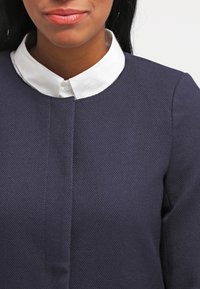 Mörkblå texturerad topp med en vit skjorta under, med en framseam och minimalistisk design.