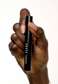 Un tube de mascara noir tenu dans une main, avec un texte blanc "BOBBI BROWN". La main a un teint naturel et des ongles bien manucurés.